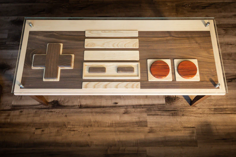 A wooden coffee table designed to resemble a classic NES controller, featuring an inlaid dark wood controller shape on the surface with lighter wood panels representing the button layout and two red circular wooden details mimicking the A and B buttons.