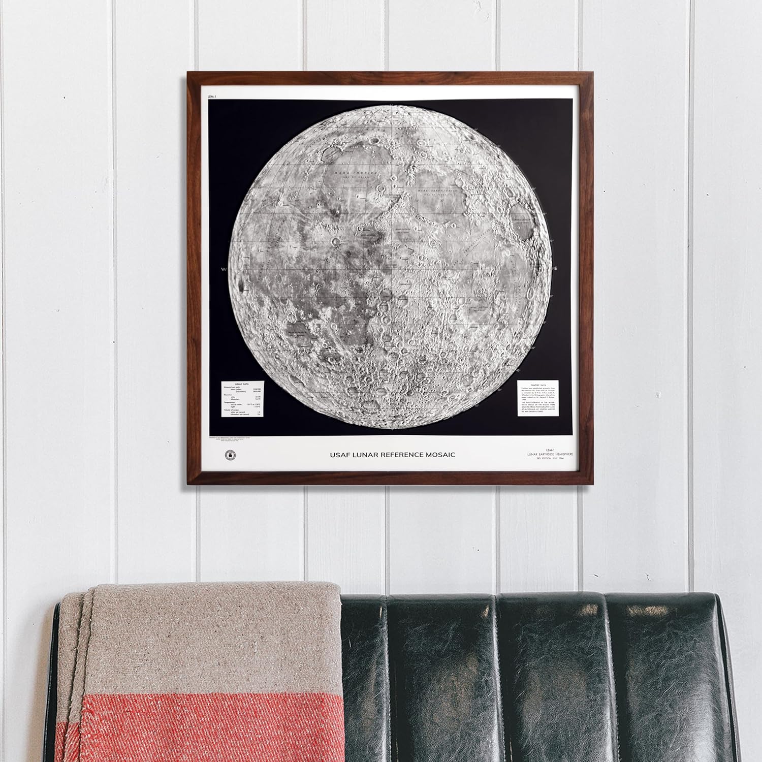A framed USAF Lunar Reference Mosaic featuring a detailed grayscale map of the moon's surface mounted in a dark wood frame, displayed on a white shiplap wall above a folded blanket and green leather ottoman.