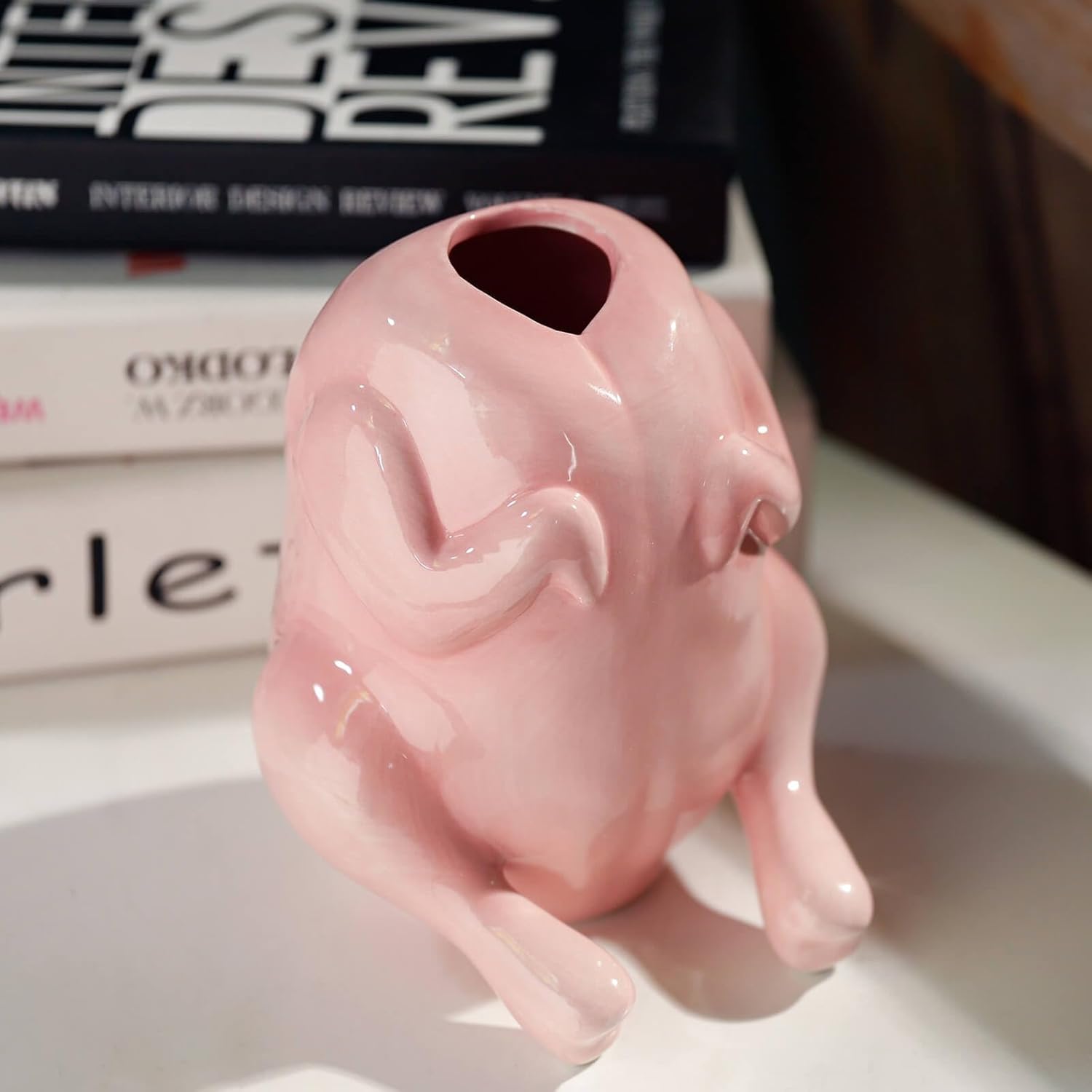 A glossy pink ceramic vase shaped like a plucked turkey, featuring a rounded body with textured feather details and an opening at the top, displayed on a white surface with books in the background.
