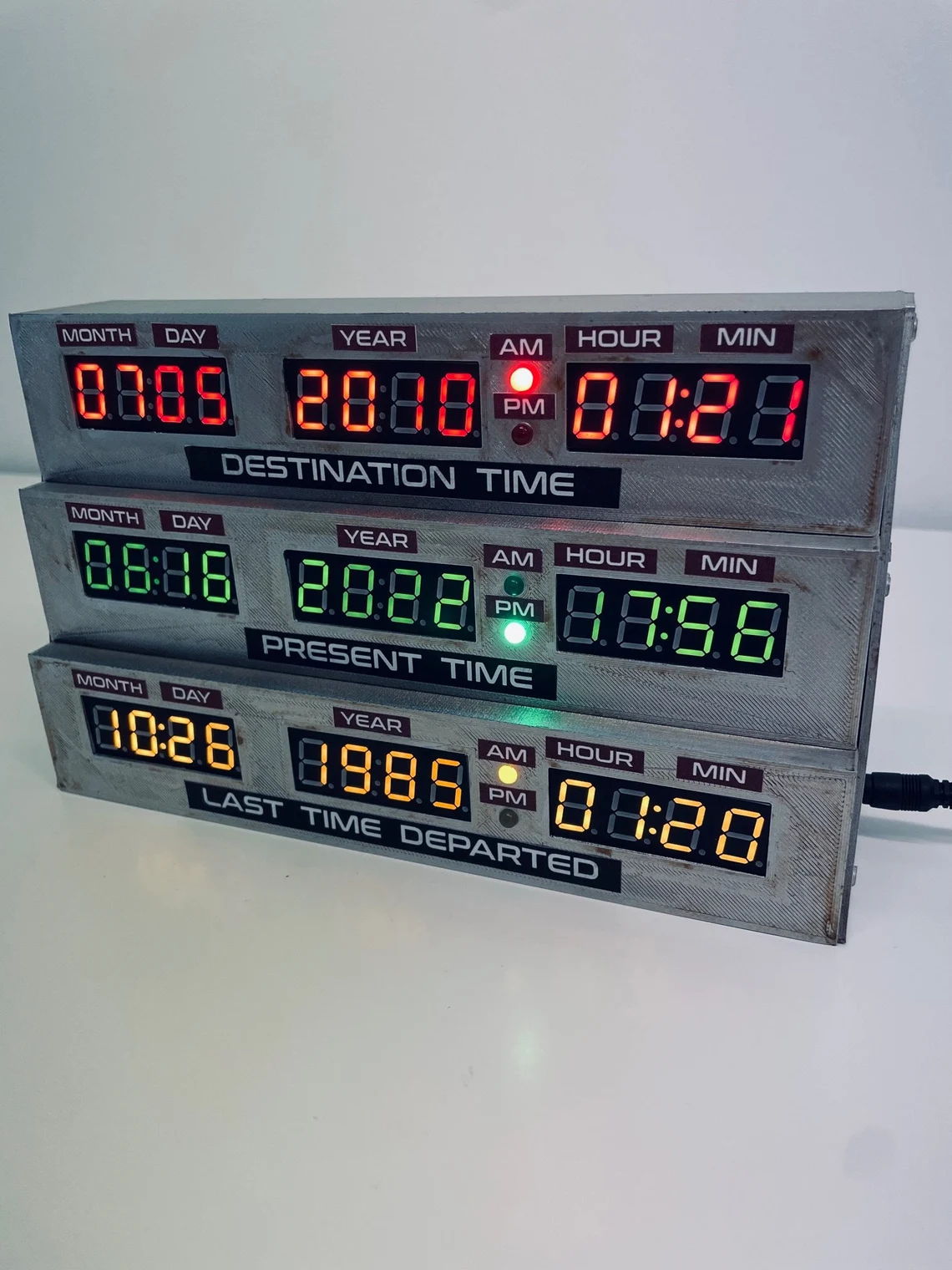 A stacked three-panel digital display clock with red, green, and yellow LED readouts showing "Destination Time," "Present Time," and "Last Time Departed" respectively, each displaying month, day, year, and time in 12-hour format with AM/PM indicators.