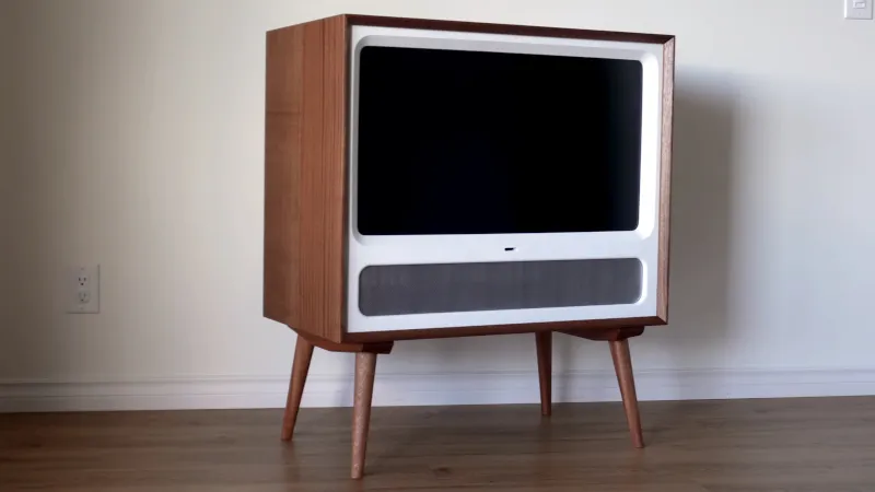 Modern TV In a Retro Case A contemporary flat-screen television integrated into a mid-century modern wooden cabinet with tapered legs, featuring a white bezel frame and speaker grille below the display.