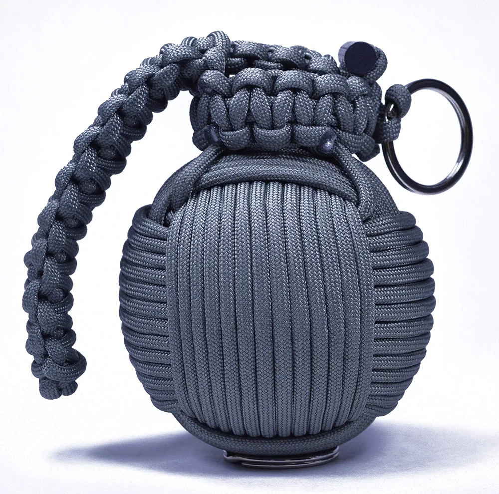 A dark gray paracord survival grenade with a spherical ribbed body, decorative knot work on top, and a braided paracord handle with a metal ring attachment, displayed against a white background.