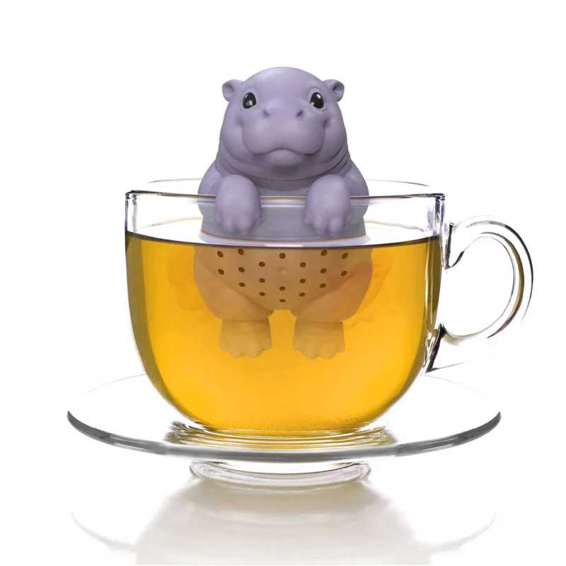 A purple silicone baby hippo tea infuser sits perched on the rim of a glass mug filled with golden tea, with the hippo's body submerged in the liquid and a matching glass saucer beneath.